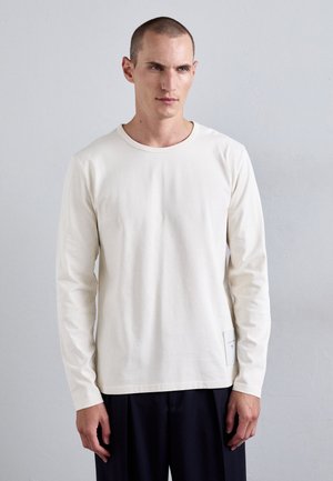 Homme portant une chemise simple à manches longues couleur blanc cassé et un pantalon foncé, se tenant contre un fond gris clair, regardant légèrement sur le côté.