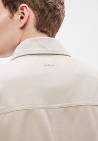 Világos bézs színű szarvasbőr dzseki gallérral, varrott részletekkel és a hát felső részén domborított "BOSS" logóval.