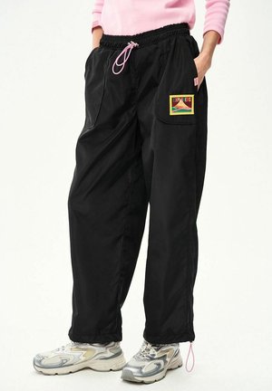 Osoba nosząca czarne, luźne spodnie z przednią naszywką z napisem "EXPLORERS" oraz szare buty sportowe.