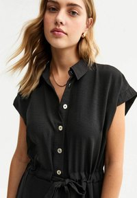 Vestido de camisa negro con botones, de manga corta, detalle de cinturón atado y tejido texturizado. Presenta botones circulares blancos a lo largo del frente.