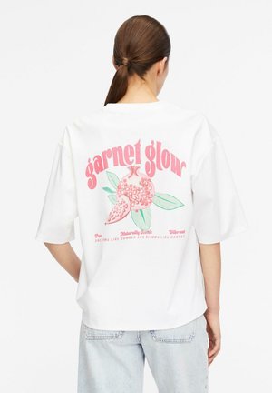 Woman with brown hair in a ponytail wears a white oversized t-shirt with pink pomegranate graphic and "garnet glow" text on the back.