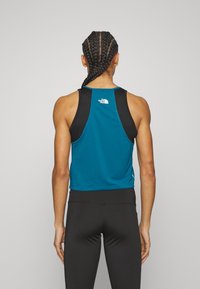 Top deportivo teal con tirantes negros, presenta una textura acanalada y un logo en la parte posterior. Combinado con leggings ajustados negros.