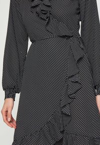 Vestido negro con lunares, de mangas largas, con detalle de volantes en el escote y el dobladillo. Hecho de una tela suave, presenta una cintura ajustada y una falda acampanada.