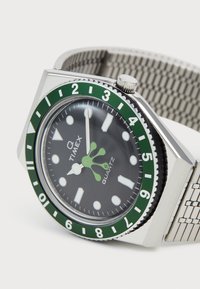 Montre-bracelet Timex avec cadran noir, lunette rotative verte marquée de 1 à 12, index horaires luminescents et bracelet en mailles métalliques argentées.