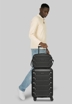 Man draagt een lichte trui en jeans en trekt een zwarte koffer met harde schaal, waarop een bijpassende handbagage tas gestapeld is.