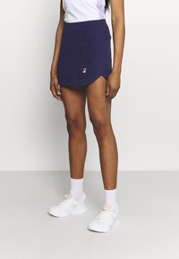 Žena oblečená v fialové sportovní sukni, bílé ponožky a bílé tenisky, stojí na jednoduchém světlem pozadí.