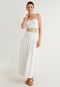 Top corto blanco con apliques florales, combinado con una falda maxi blanca con cintura fruncida. Lleva sandalias marrones de tiras.