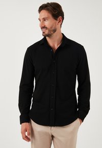 Camisa negra de manga larga con botones, confeccionada en una tela suave, que presenta un cuello clásico y un diseño entallado, con botones negros en la parte frontal.