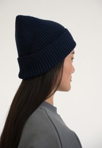 Gorro de punto azul marino con textura acanalada y parte superior puntiaguda. Usado con una sudadera gris, que destaca su ajuste ceñido y diseño informal.