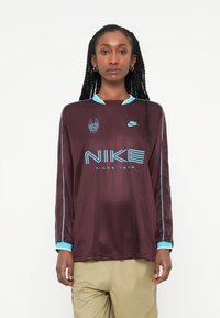 Dolga majica Nike v temno rdeči barvi z svetlo modrimi detajli, opremljena z rebrastim ovratnikom, logotipom in boldnim napisom "NIKE" spredaj.