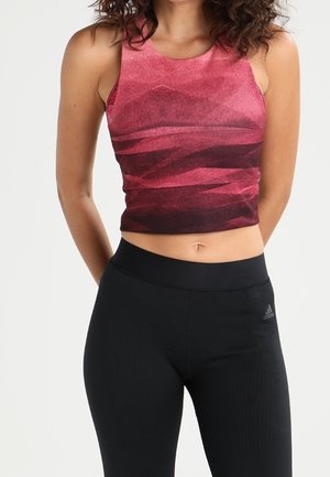 Femme portant un crop top rouge sans manches à motifs et un legging de sport noir taille haute avec un logo Adidas sur la hanche.