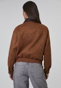 Kobieta z długimi ciemnymi włosami nosi brązową kurtkę z zamszu z elastycznym paskiem w talii oraz szare jeansy z wysokim stanem, zwrócona tyłem na jednolitym tle.