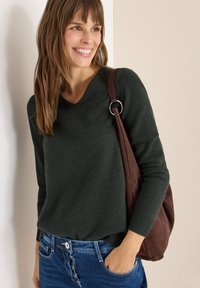 Dunkelgrüner Wollpullover mit V-Ausschnitt, langen Ärmeln und gerippten Bündchen. Kombiniert mit blauen Jeans und einer braunen Wildleder-Schultertasche.