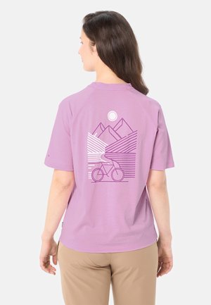 Frau mit langen braunen Haaren, die ein lavendelfarbenes T-Shirt mit einem Motiv aus Berg, Sonne und Fahrrad auf dem Rücken trägt, kombiniert mit beigefarbenen Hosen.