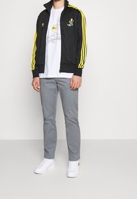 Svart zip-up jacka med gula ränder och sidofickor. Har en tecknad karaktär på bröstet. Bärs med grå byxor och vita skor.