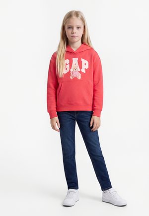 Jong meisje met lang blond haar draagt een rode GAP-hoodie met een teddybeerprint, blauwe spijkerbroek en witte sneakers, staand tegen een egale achtergrond.