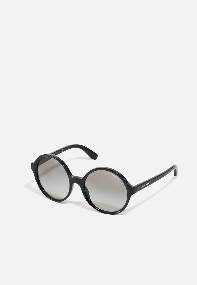 Óculos de sol redondos pretos com um acabamento brilhante, lentes cinzentas em degradê e hastes texturizadas que exibem um logótipo. Construção leve em plástico.