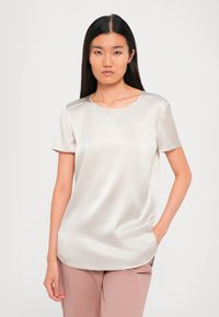 Donna con capelli lunghi e scuri, che indossa una camicia beige lucida a maniche corte e pantaloni leggermente malva, in piedi con una mano in tasca su sfondo bianco.