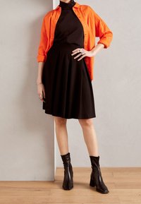 Oranžová košile s knoflíky přeložená přes černý rolák, v kombinaci s černou plisovanou sukní a černými kotníkovými botami na širokém podpatku.