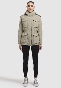Khaki gewatteerde jas met capuchon, voorzien van vier voorzakken en een trekkoord in de taille, gecombineerd met zwarte leggings en hoge sneakers.