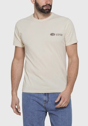 Uomo con indosso una t-shirt beige a maniche corte con il logo "CARRERA BLUE AGE" sul petto, abbinata a jeans blu in denim, in piedi su uno sfondo uniforme.