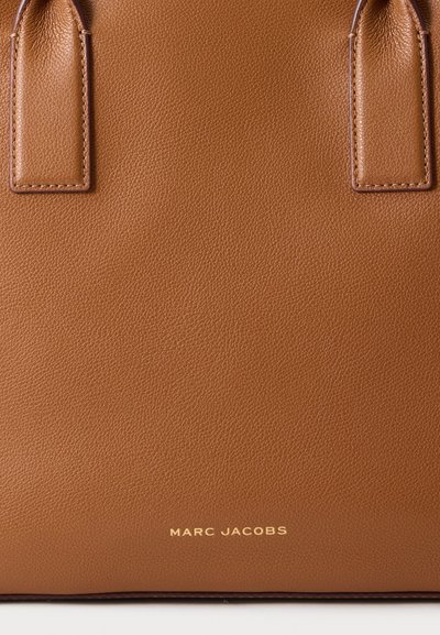 Sac à main en cuir marron avec deux poignées cousues et un logo Marc Jacobs doré centré près du bas.
