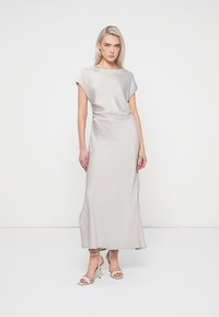 Μακριά φόρεμα από ανοιχτό γκρι ύφασμα, με χαλαρή σιλουέτα, κοντά μανίκια και διακριτική γυαλάδα. Συνδυασμένο με μπεζ σανδάλια με τακούνι.