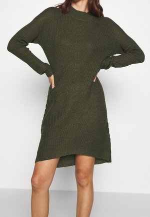 Femme portant une robe-pull en maille vert foncé à manches longues, debout, les mains sur les hanches, devant un fond uni.