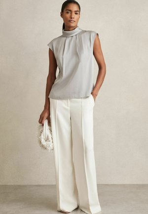 Gray sleeveless top with a high collar and pleats, paired with white wide-leg trousers. Model holds a fluffy white handbag.