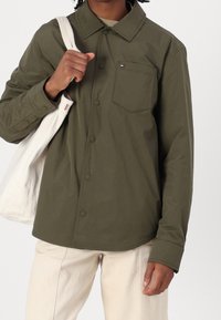 Olivengrøn skjorte med knapper i let vævet stof, med en brystlomme og trykknapper. Kombineret med cremefarvede bukser og en hvid tote-taske.