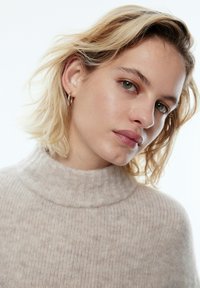 Beige gestrickter Rollkragenpullover mit geripptem Muster und weicher Textur. Das Model hat kurze, wellige blonde Haare und silberne Creolen.