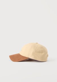 Μπεζ καπέλο του μπέιζμπολ με καφέ κυρτή γείσο, που βρίσκεται πάνω σε λευκή επιφάνεια, θέα από το πλάι.