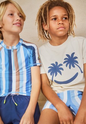 Δύο αγόρια καθισμένα δίπλα-δίπλα, το ένα με μπλε ριγέ μαγιό και το άλλο με μπεζ μπλούζα που έχει χαρούμενη παλάμη και αντίστοιχες ριγέ βερμούδες.
