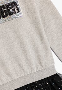 Felpa grigia con la scritta in paillettes "BIGGER", presenta un orlo a coste e una gonna nera in tulle con motivi a stelle multicolori.