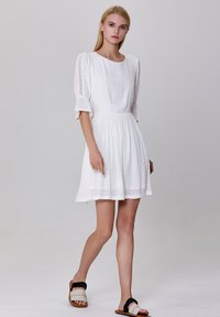 Robe blanche mi-longue en tissu texturé, avec de courtes manches bouffantes et des accents en dentelle. Associée à des sandales plates avec des bordures noires.