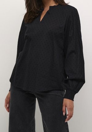 Femme portant un chemisier noir texturé à manches longues avec un petit col en V et des poignets élastiques, associé à un jean gris foncé.
