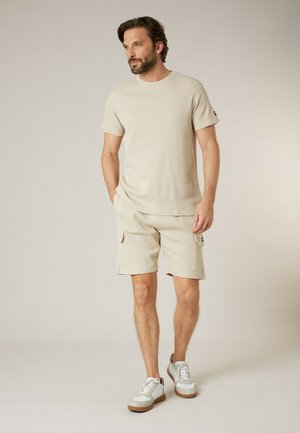 Homme barbu portant une chemise à manches courtes beige texturée, un short cargo et des baskets blanches, debout les mains dans les poches.