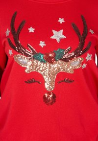 Rode trui met daarop een versierd rendiergezicht met pailletten, steraccenten en hulstdetails, met nadruk op textuur en levendige kleuren.