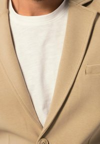 Béžový blazer s klopami a jedním knoflíkem, položený na strukturovaném bílém tričku, které ukazuje čistý a jednoduchý design.