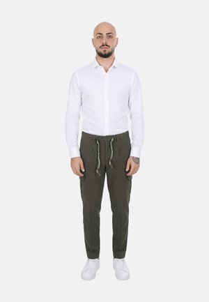 Uomo calvo con barba in piedi di fronte, indossa una camicia bianca con bottoni, pantaloni verde oliva con coulisse, sneakers bianche e un tatuaggio al polso.