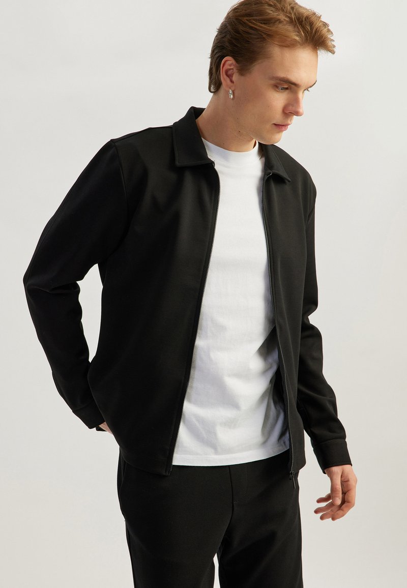 Black zip-up jacket with a collared neckline and long sleeves, made from smooth fabric, worn over a white T-shirt. Minimalist design.