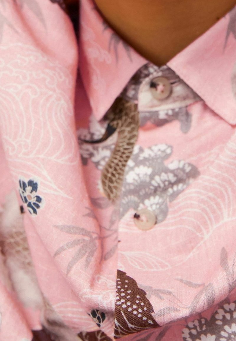 Gros plan sur une chemise rose à boutons avec des motifs floraux et de feuilles en tons marron, gris et bleu sur un tissu usé.