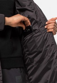 Zwarte geïsoleerde jas met een textuurvoering, met een smartphonevak aan de binnenkant van de linkerkant. Hand die een telefoon in het vak steekt.