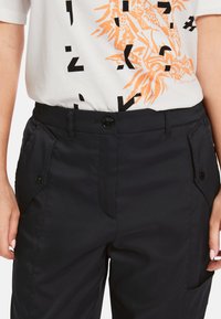 Schwarze Hosen mit einer glatten Textur, ausgestattet mit einem Knopfverschluss, Fronttaschen und einem flachen Bund, kombiniert mit einem grafischen T-Shirt in Weiß und Orange.