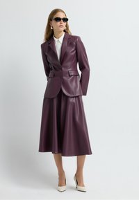 Blazer en simili cuir violet foncé avec une coupe ajustée, associé à une jupe évasée assortie. Une chemise blanche à col et des lunettes de soleil noires complètent le look.