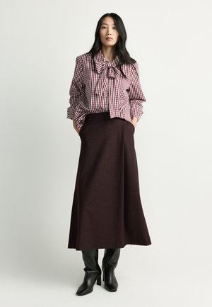 Woman wearing a red and white checkered blouse with a bow, a dark brown midi skirt, and black heeled boots standing against a plain background.