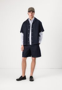 Denim antrekk med kortermet skjorte, knappelukking foran og matchende shorts. Skjorten har lange, lyseblå stribede ermer. Svarte loafere og en rutete caps fullfører looket.