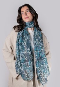 Blomsterhalsedisse i teal- og blåtoner, lavet af letvægtigt stof med et delikat mønster, hænger løst om halsen, suppleret af en beige frakke.