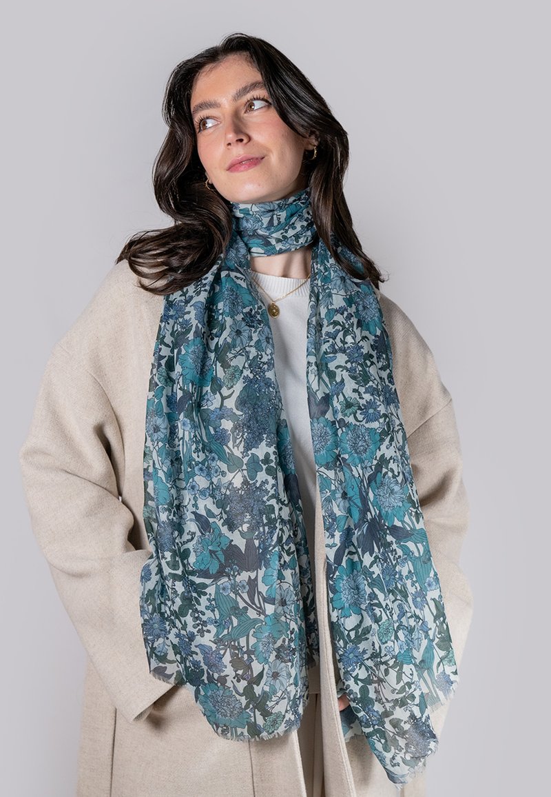 Blomsterhalsedisse i teal- og blåtoner, lavet af letvægtigt stof med et delikat mønster, hænger løst om halsen, suppleret af en beige frakke.