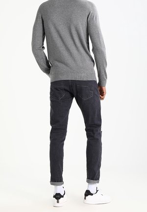 Mężczyzna ubrany w szary sweter, ciemne podwinięte dżinsy, białe skarpetki i białe trampki stoi tyłem do przodu.